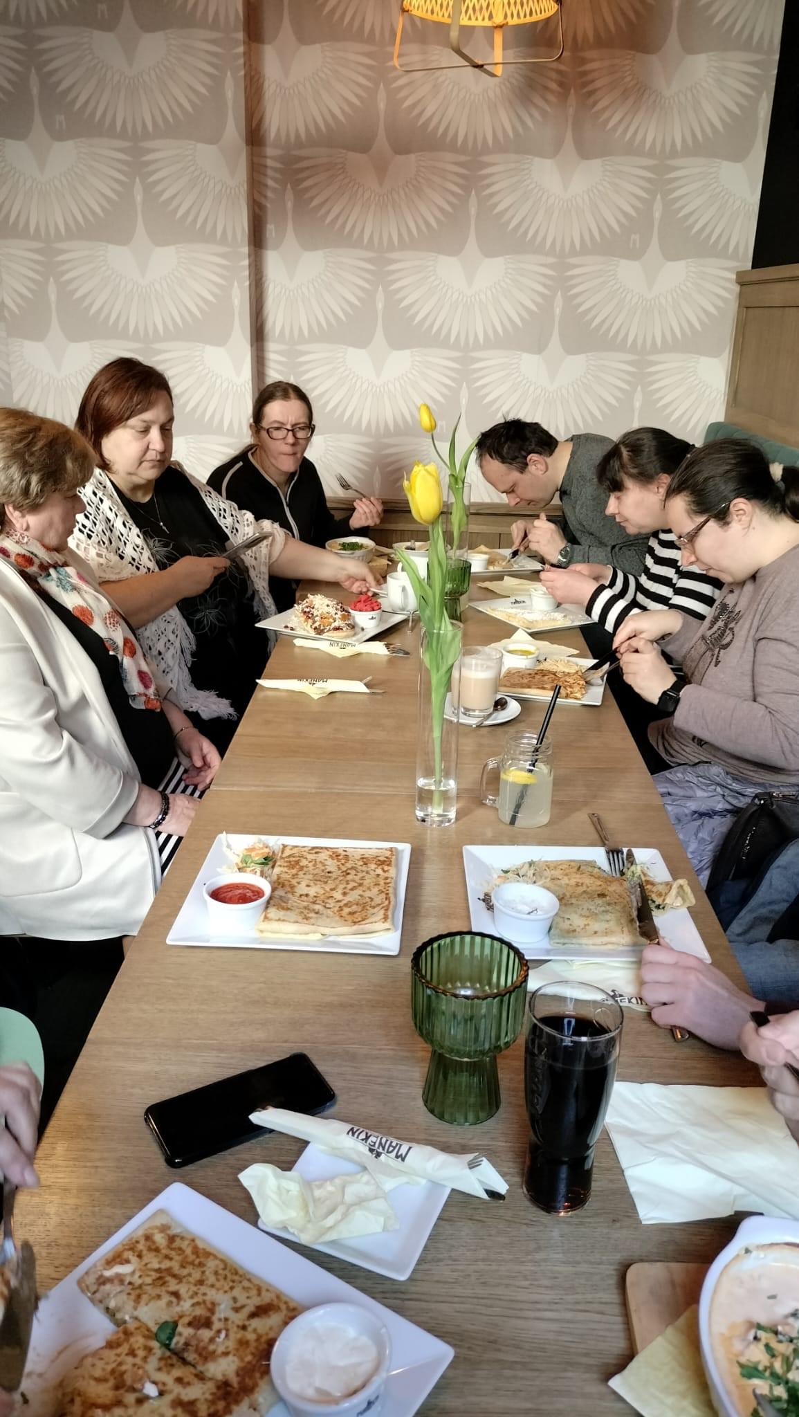 Grupa osób przy stole podczas konsumpcji posiłków. Na stole jedzenie oraz kwiaty. 