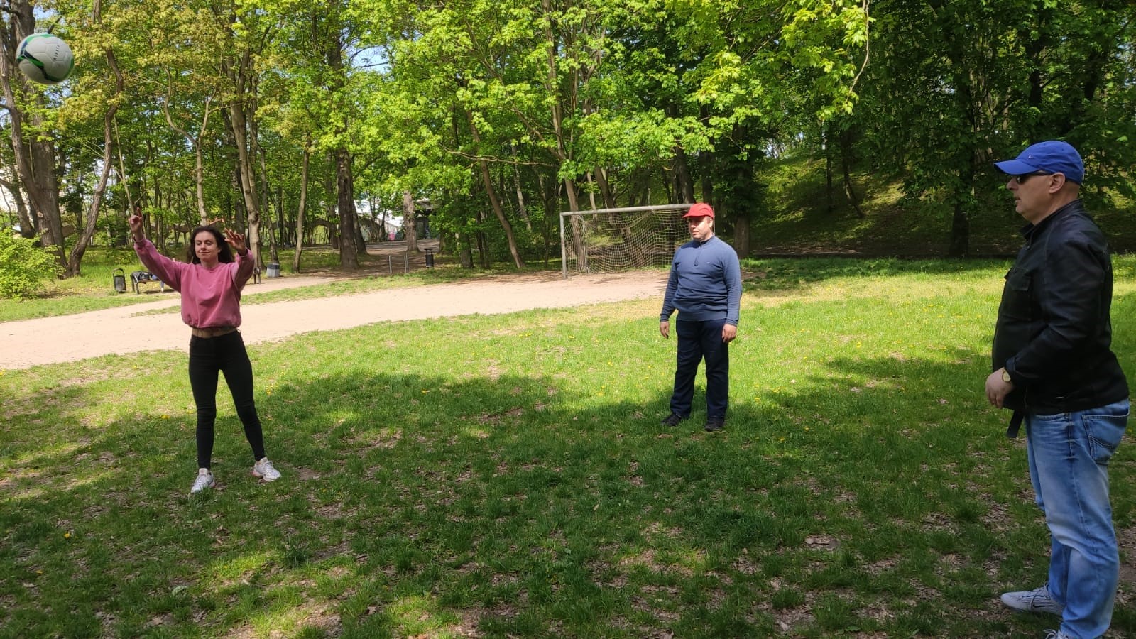 Kobieta i dwóch mężczyzn na polanie w otoczeniu drzew odbijają piłkę do gry w siatkówkę. 