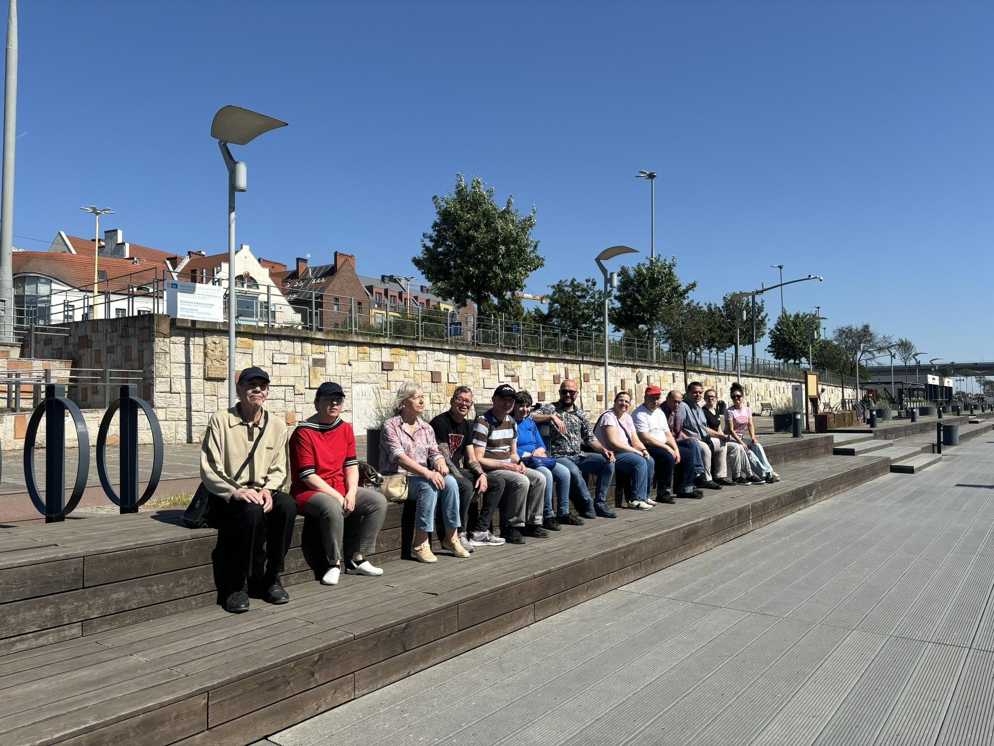 Grupa osób siedząca na bulwarach, w tle zabudowa miejska oraz zieleń. 