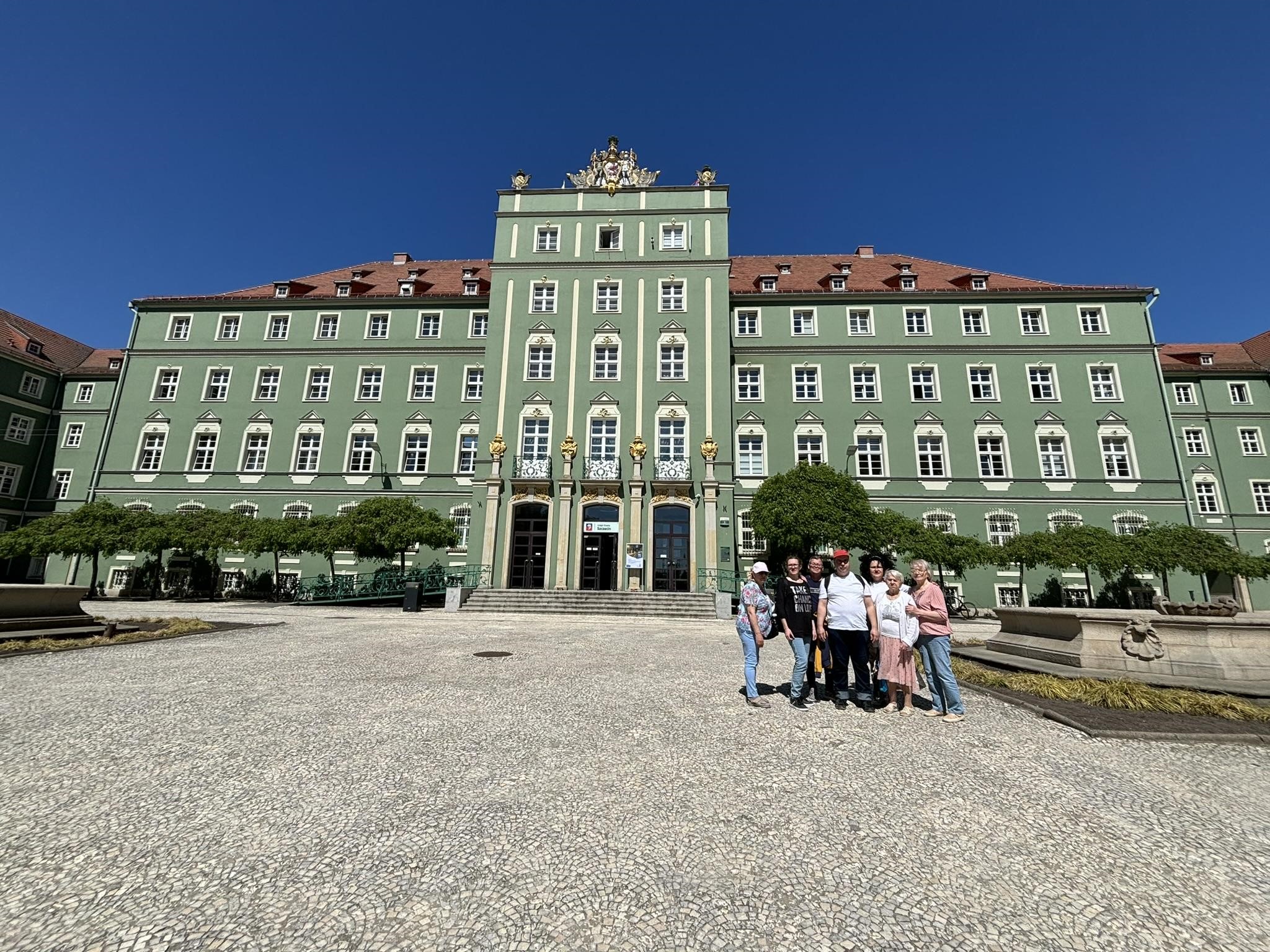 Grupa osób pozująca do zdjęcia na tle budynku Urzędu Miasta Szczecin, koloru zielonego. 