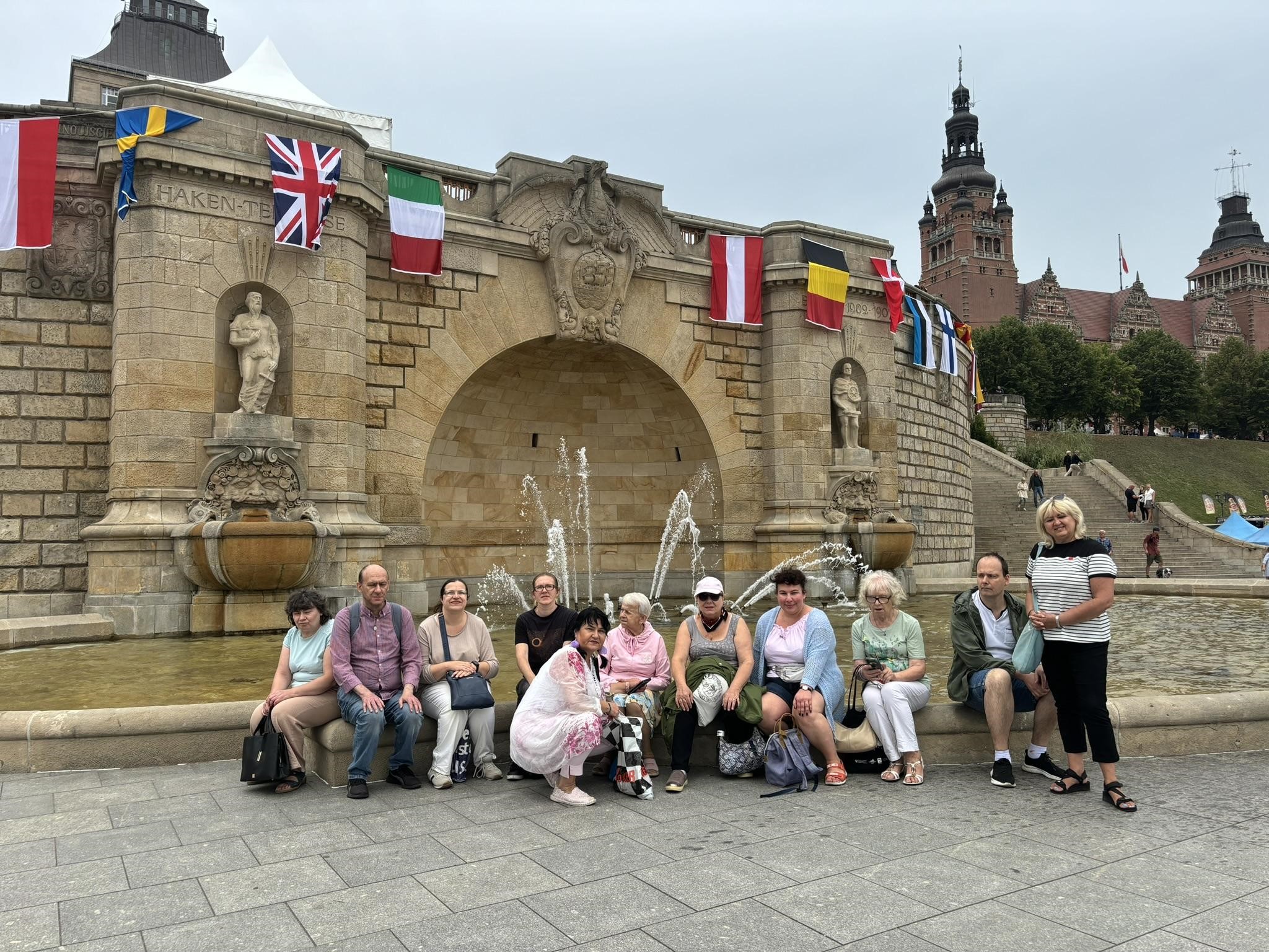 : Grupa osób pozująca na tle fontanny na Wałach Chrobrego w Szczecinie. W tle wywieszone kolorowe flagi. 