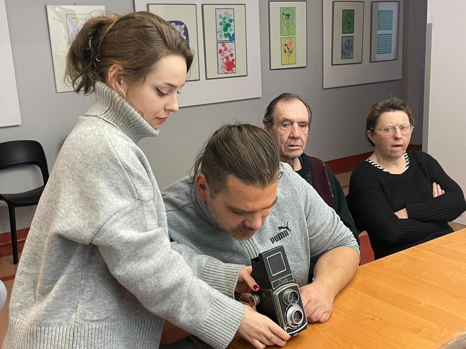 Kobieta trzymająca lampę. Przed kobietą znajduje się stół, na nim leży sprzęt fotograficzny. W tle widać rozwinięty ekran. Dookoła stołu siedzą ludzie.