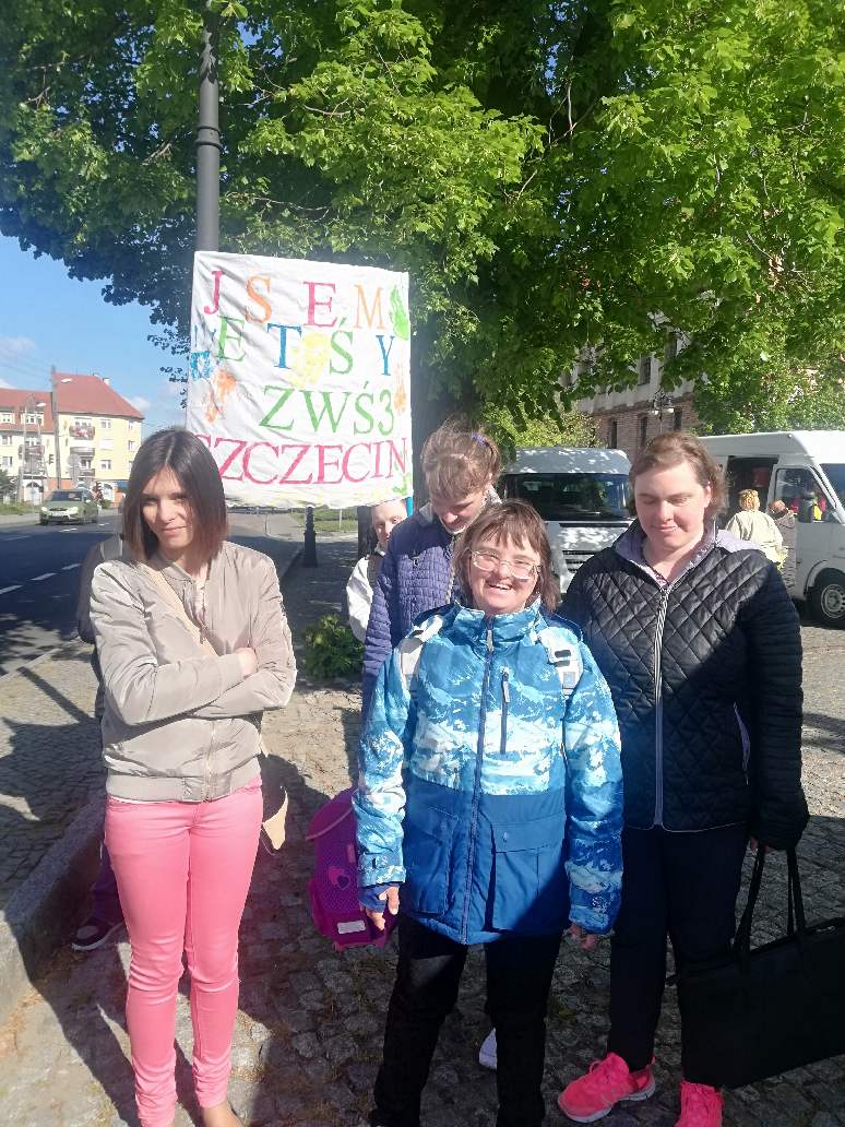 Cztery dziewczyny stoją na chodniku. Za nimi znajduje się plakat, który jest trzymany przez chłopaka. 