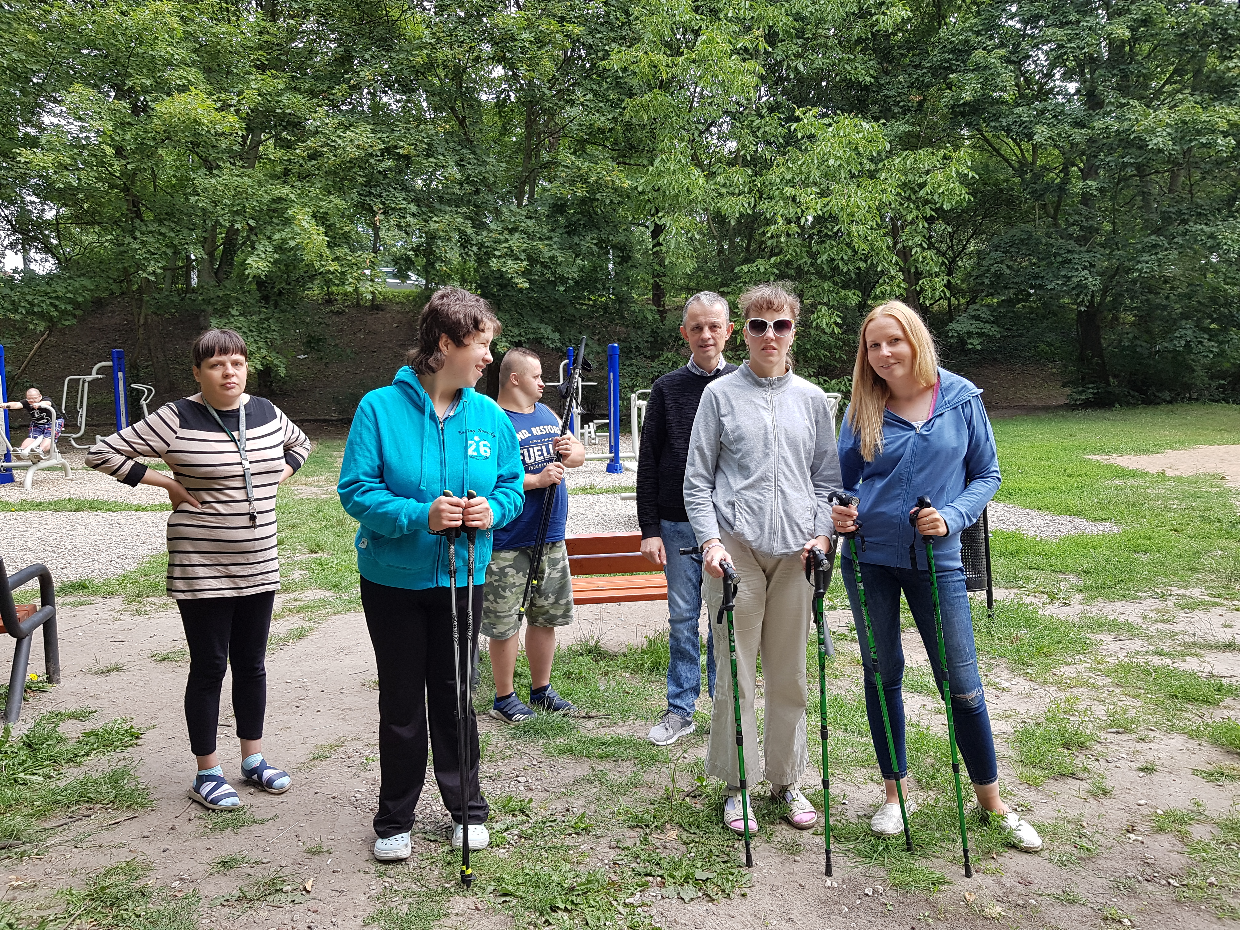 Pięciu uczestników wraz z terapeutką stoją w parku. W rękach trzymają kije do nordic walking. 