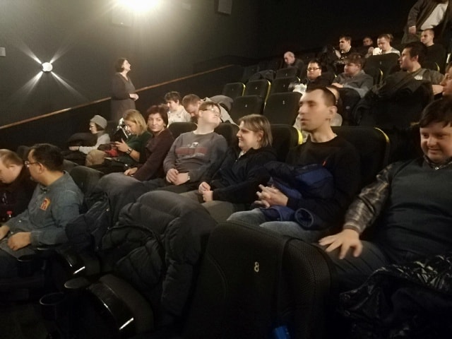 W sali kinowej grupa uczestników siedzi w fotelach. W tle widać innych ludzi.