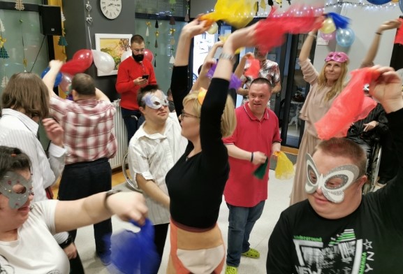Grupa ludzi ustawionych w rozsypce stoi z uniesionymi rękami trzymając w nich kolorowe chusty. 3 osoby mają założone na twarzach ozdobne maski.
