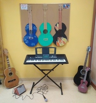 Na środku stoi keyboard. Nad nim zawieszone są tekturowe gitary, poniżej na podłodze leży mikrofon i głośnik, a na bokach stoją gitary, ukulele oraz wzmacniacz. 