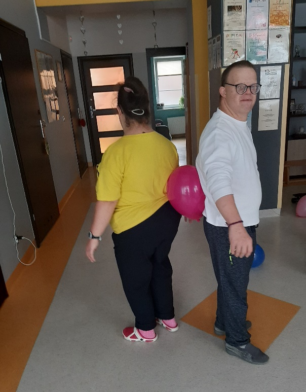 Dwie osoby stoją do siebie plecami trzymając ,między sobą balon.