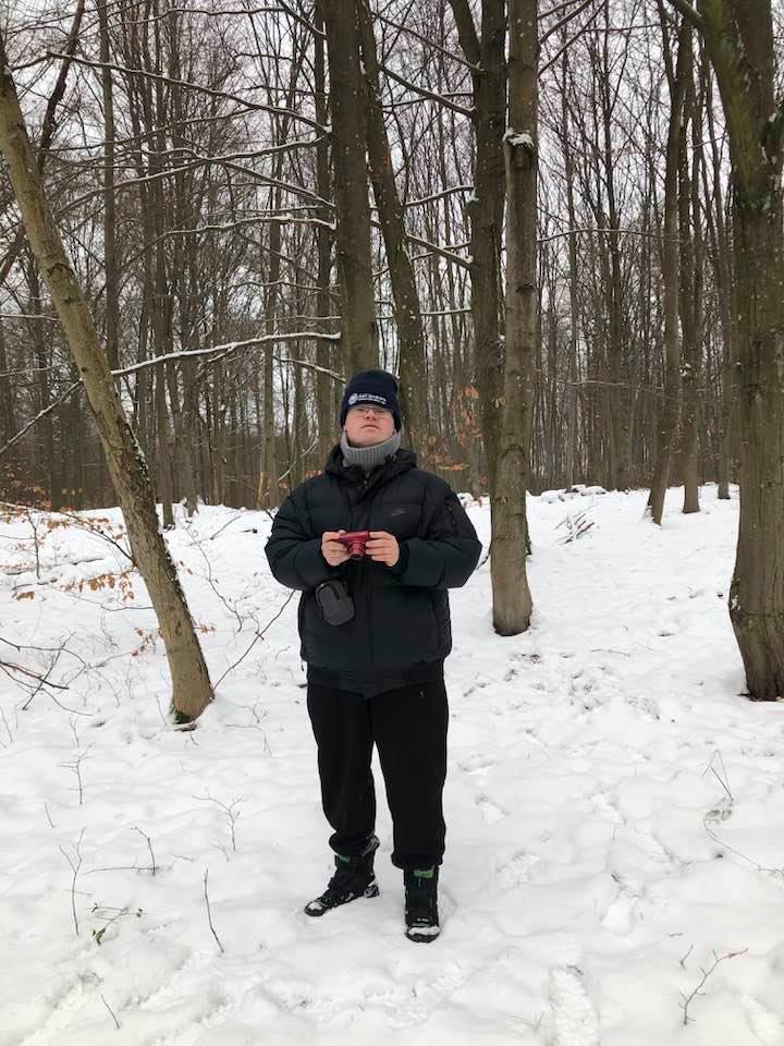 Mężczyzna stoi na śniegu, trzyma w rękach aparat fotograficzny. Z każdej strony otaczają go drzewa. 