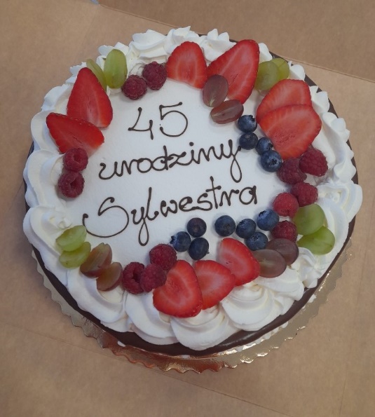 Na stole leży tort ozdobiony owocami, z napisem 45 urodziny Sylwestra.