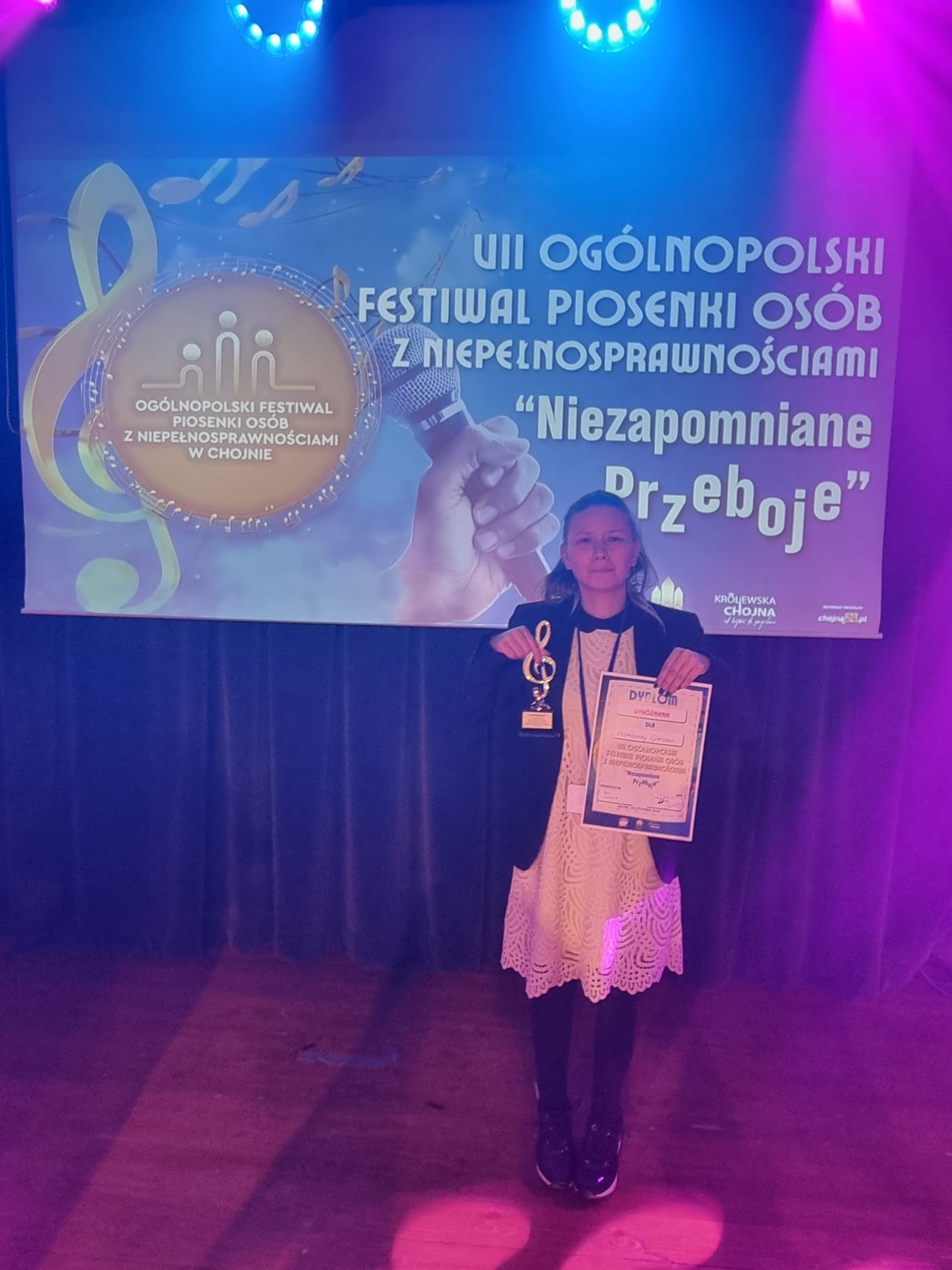 Kobieta pozuje do zdjęcia. W prawej ręce trzyma statuetkę a w lewej dyplom. W tle duży baner.