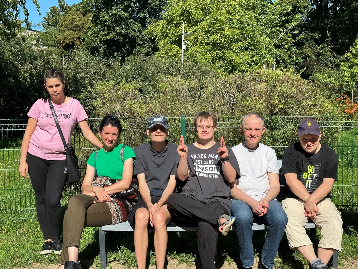 Jedna kobieta stoi przy ławce. Jedna kobieta i czterech mężczyzn siedzi na ławce. W tle drzewa.