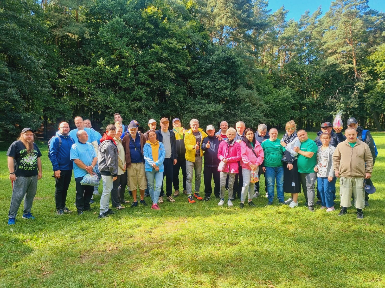 Grupa kobiet i mężczyzn pozuje na trawie do zdjęcia. W tle drzewa. 