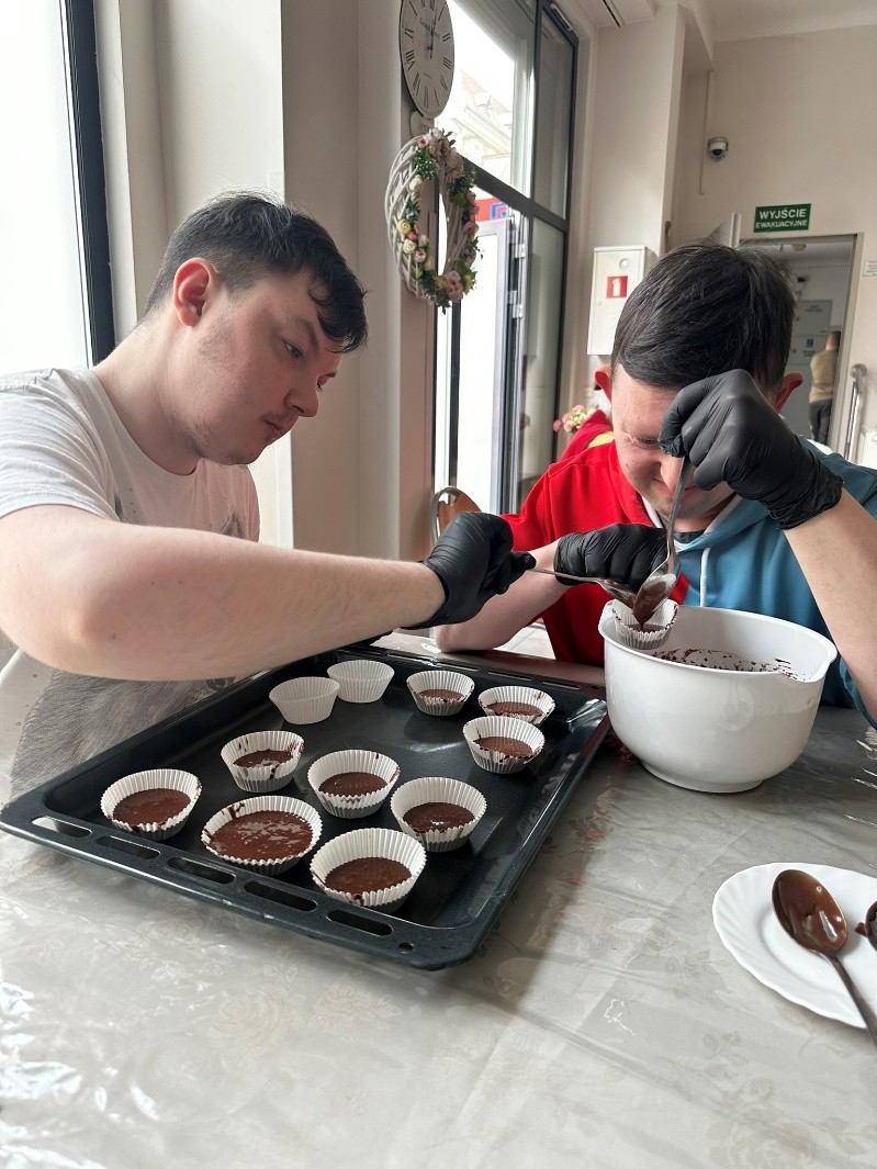 Dwóch mężczyzn przygotowuje babeczki do wypieku. W rękach trzymają łyżki. Nakładają masę z miski do formy. Na stole znajduje się blacha do pieczenia a na niej formy wypełnione masą do pieczenia.