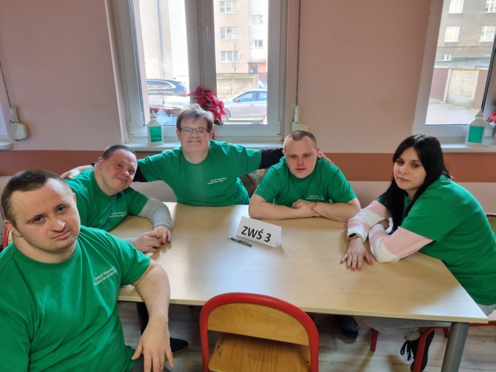 Czterech mężczyzn i jedna kobieta siedzą blisko siebie patrząc w obiektyw. Wszyscy mają na sobie zielone koszulki na krótki rękaw. 