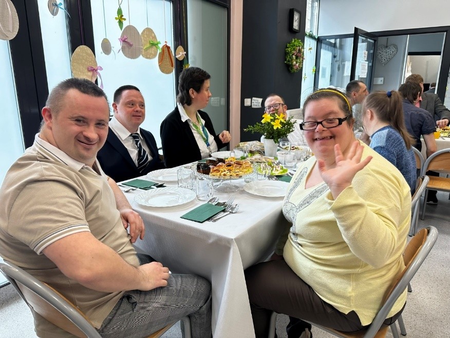 Na pierwszym planie kobieta i mężczyzna uśmiechają się, patrzą prosto w obiektyw a kobieta macha. Na stole obrus, zastawa i potrawy. W tle kobiety i mężczyźni.